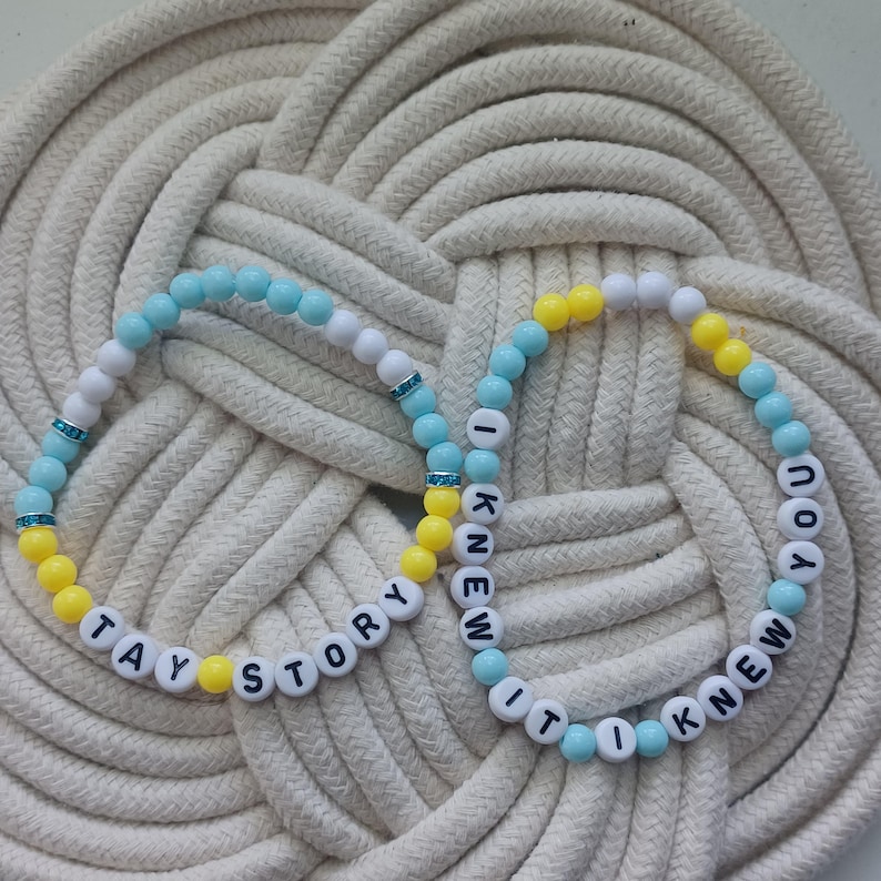 Puede incluir: Dos pulseras el&aacute;sticas de cuentas con abalorios redondos blancos, azul claro y amarillos. Una pulsera muestra el texto negro TAY STORY, mientras que la otra muestra el texto negro I KNEW IT I KNEW YOU. Ambas pulseras incluyen peque&ntilde;os detalles azules.