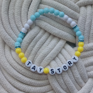 Puede incluir: Una pulsera de cuentas con una secuencia de cuentas redondas de color azul claro, blanco y amarillo. La pulsera incluye peque&ntilde;os espaciadores plateados con pedrer&iacute;a azul y cuentas con letras blancas que forman la frase TAY STORY en negro.