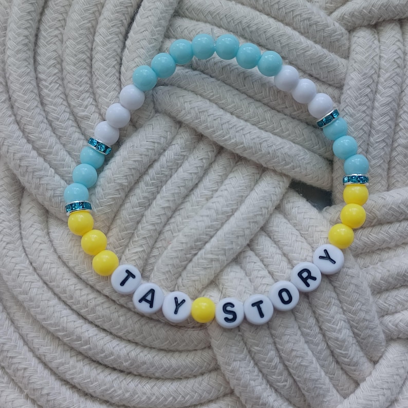 Puede incluir: Una pulsera de cuentas con una secuencia de cuentas redondas de color azul claro, blanco y amarillo. La pulsera incluye peque&ntilde;os espaciadores plateados con pedrer&iacute;a azul y cuentas con letras blancas que forman la frase TAY STORY en negro.