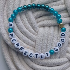 May include: A beaded bracelet with the words "PERFECTLY GOOD" spelled out in white letter beads. The bracelet features teal and dark teal beads, with a small gold heart charm. The bracelet is displayed on a white rope surface.