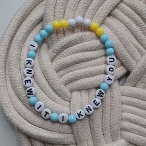 Puede incluir: Una pulsera circular de cuentas con esferas blancas, azul claro y amarillas. Las cuentas blancas tienen letras negras impresas que forman la frase I KNEW IT I KNEW YOU, dispuestas en una secuencia repetitiva alrededor de la pulsera.