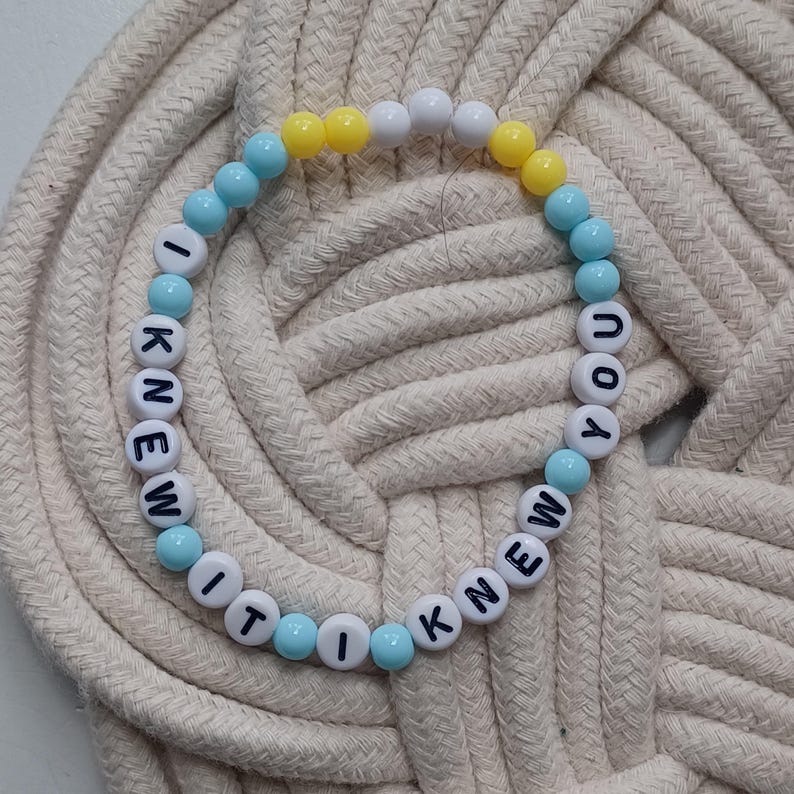 Puede incluir: Una pulsera circular de cuentas con esferas blancas, azul claro y amarillas. Las cuentas blancas tienen letras negras impresas que forman la frase I KNEW IT I KNEW YOU, dispuestas en una secuencia repetitiva alrededor de la pulsera.