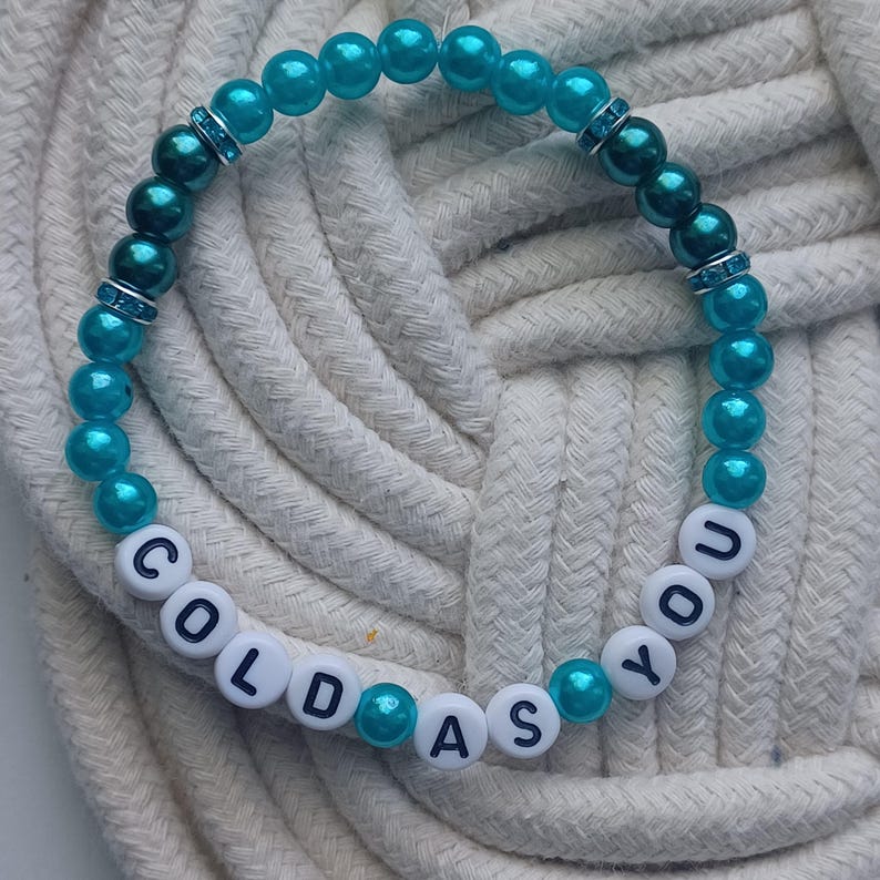 May include: A beaded bracelet with turquoise and dark teal beads. The bracelet features white letter beads spelling "COLD AS YOU". The bracelet is displayed on a white rope surface.