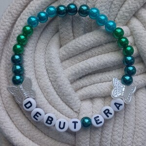 May include: A beaded bracelet with teal, green, and blue beads. The bracelet features the word "DEBUTERA" spelled out in white letter beads and two silver butterfly charms. The bracelet is displayed on a white rope surface.