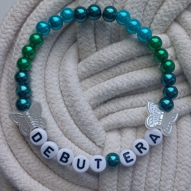 May include: A beaded bracelet with teal, green, and blue beads. The bracelet features the word "DEBUTERA" spelled out in white letter beads and two silver butterfly charms. The bracelet is displayed on a white rope surface.