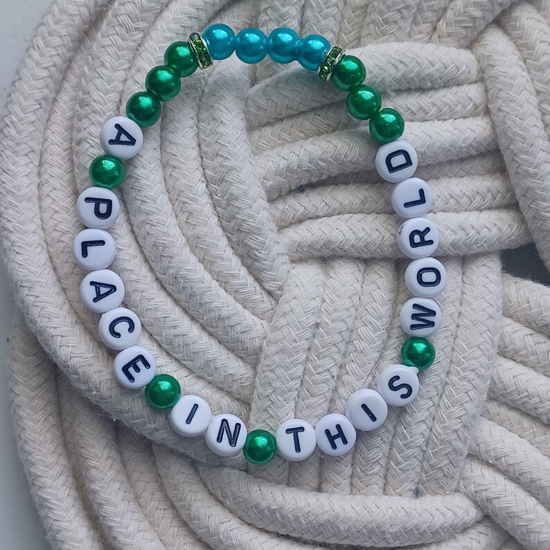 May include: A beaded bracelet with the words "A PLACE IN THIS WORLD". The bracelet features white letter beads, green and teal beads, and is set against a white rope background. The bracelet is a piece of jewelry.
