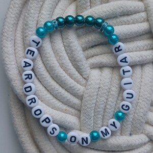 May include: A beaded bracelet with the words "TEARDROPS ON MY GUITAR" spelled out in white letter beads. The bracelet features turquoise and dark teal beads, with a few clear rhinestone accents. The bracelet is displayed on a white rope surface.