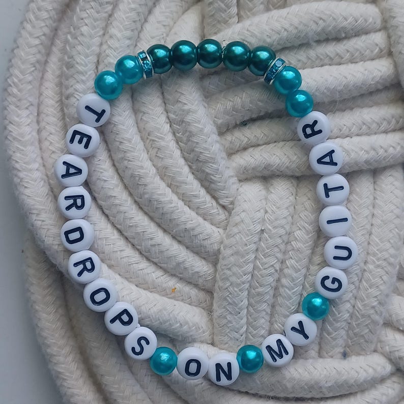 May include: A beaded bracelet with the words "TEARDROPS ON MY GUITAR" spelled out in white letter beads. The bracelet features turquoise and dark teal beads, with a few clear rhinestone accents. The bracelet is displayed on a white rope surface.