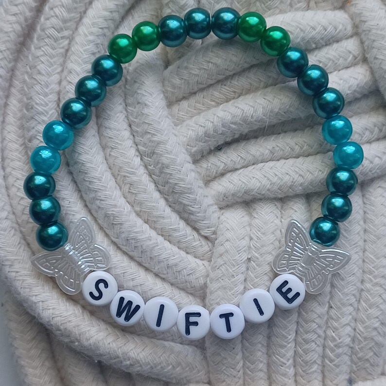 May include: A beaded bracelet with a gradient of teal and green beads. The bracelet features the word "SWIFTIE" spelled out in white letter beads and two butterfly charms.