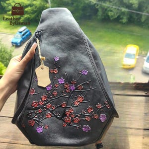 Puede incluir: Bolso bandolera de cuero negro con diseño floral en relieve en rojo y morado. El bolso tiene cierre de cremallera y una pequeña etiqueta. El bolso está diseñado para llevarse sobre un hombro.