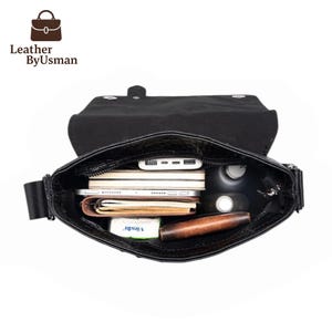 K&ouml;nnte beinhalten: Schwarze Ledertasche mit offenem Deckel, die den Inhalt zeigt: B&uuml;cher, Geldb&ouml;rse, Telefon, Flasche und andere Gegenst&auml;nde. Die Tasche hat einen Schulterriemen. Der Text "Leather by Usman" ist sichtbar.