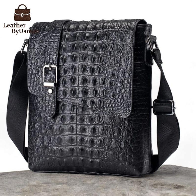 K&ouml;nnte beinhalten: Schwarze Leder-Umh&auml;ngetasche mit Krokodilhautstruktur. Die Tasche hat einen Klappenverschluss mit silberner Schnalle und einen schwarzen, verstellbaren Schulterriemen. Der Text "Leather By Usn" befindet sich oben links auf der Tasche.