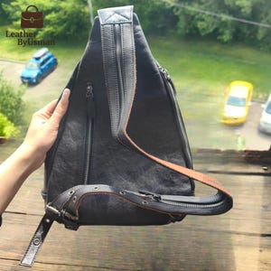May include: Black leather sling bag with a brown strap and silver zippers. The bag has a unique triangular shape and a single shoulder strap. The bag is held by a person's hand. The bag has the text "Leather By Usman".