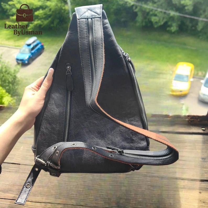 May include: Black leather sling bag with a brown strap and silver zippers. The bag has a unique triangular shape and a single shoulder strap. The bag is held by a person's hand. The bag has the text "Leather By Usman".