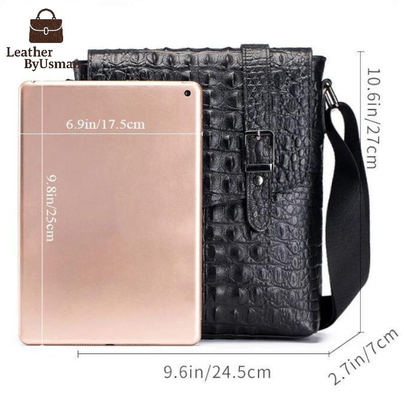 K&ouml;nnte beinhalten: Schwarze Leder-Umh&auml;ngetasche mit Krokodiltextur. Die Tasche ist 24,5 cm breit, 27 cm hoch und 7 cm tief. Ein Tablet, 17,5 cm breit und 25 cm hoch, wird zum Gr&ouml;&szlig;envergleich neben die Tasche gelegt. Die Tasche hat einen Schnallenverschluss.