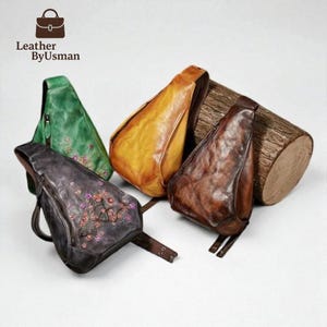 May include: Four leather sling bags in shades of green, yellow, brown, and dark gray, with a wooden log in the background. The dark gray bag features floral embroidery. The bags have adjustable straps and zippers. The logo "Leather by Usman" is in the top left corner.