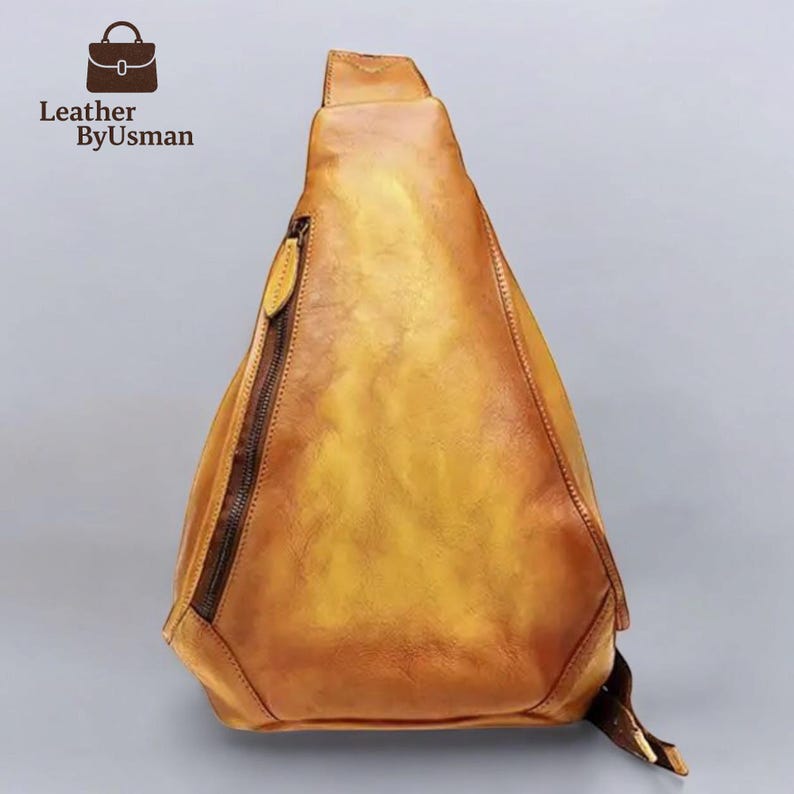 May include: A yellow-brown leather sling bag with a diagonal zipper and a strap. The bag has a triangular shape and a textured surface. The logo "Leather by Usman" is in the upper left corner.