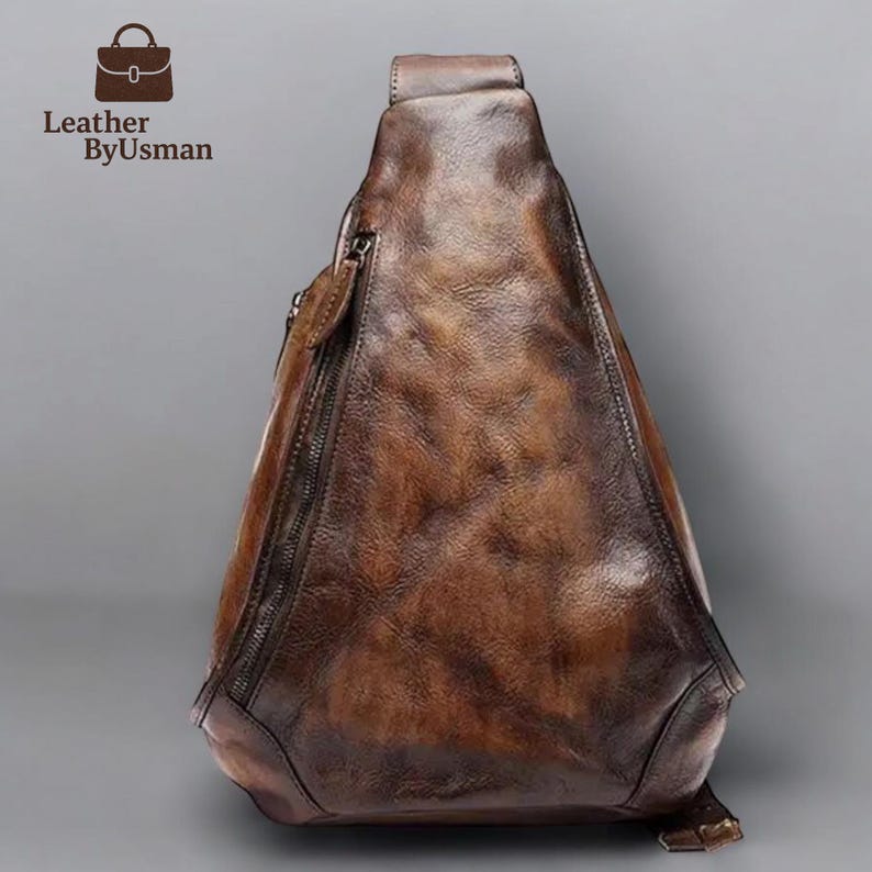 May include: A brown leather sling bag with a textured finish and a zipper closure. The bag has a triangular shape and a strap that goes over the shoulder. The text "Leather ByUsman" is in the upper left corner.