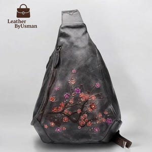 May include: A gray leather sling bag with a floral design in red and purple. The bag has a zipper closure and a brown strap. The brand name "Leather ByUsman" is visible in the upper left corner.