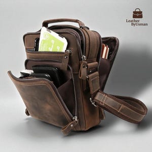 May include: A brown leather messenger bag with multiple zippered compartments and a top handle. The bag has a shoulder strap and is filled with items, including a tablet and a phone. The bag has a distressed finish and visible stitching.