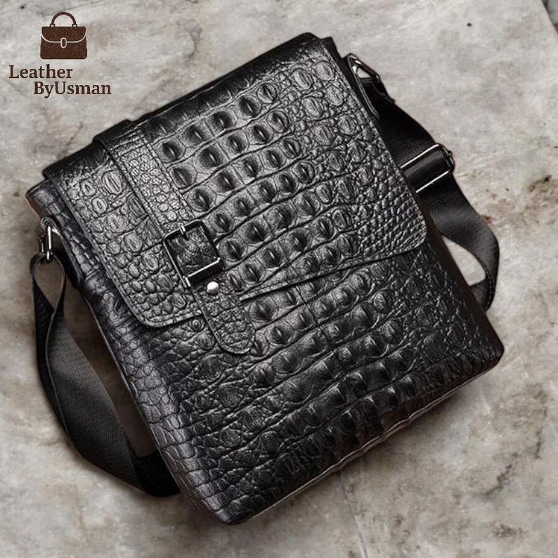 K&ouml;nnte beinhalten: Schwarze Leder-Umh&auml;ngetasche mit gepr&auml;gter Krokodil-Optik. Sie hat einen Klappverschluss mit silberner Schnalle und einen verstellbaren Schulterriemen. Die rechteckige Tasche hat ein modernes Design.