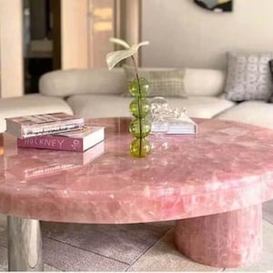 May include: A round, pink quartz coffee table with a polished surface. The table has a cylindrical base and chrome legs. Two books are stacked on top, and a green glass vase with a white flower is on the table.