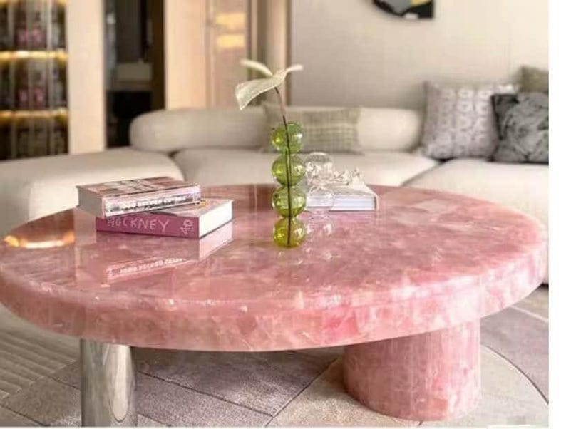 May include: A round, pink quartz coffee table with a polished surface. The table has a cylindrical base and chrome legs. Two books are stacked on top, and a green glass vase with a white flower is on the table.