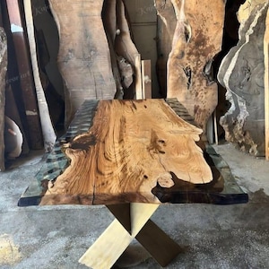 Puede incluir: Una mesa de madera hecha a mano con una tapa de vetas de madera natural y un borde de resina epoxi transparente. La mesa está soportada por una base dorada en forma de X. Las dimensiones de la mesa no son visibles en la imagen.