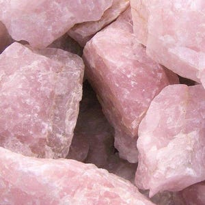 May include: A collection of raw rose quartz crystals in varying sizes. The stones have a soft, opaque pink hue and a rough, textured surface. The crystals are clustered together, showcasing their natural, unpolished state.