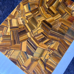 May include: A rectangular tabletop with a mosaic design made of tiger's eye gemstone. The surface features a warm palette of golden brown, amber, and dark brown tones, with visible natural patterns. The tabletop is set against a light blue surface.