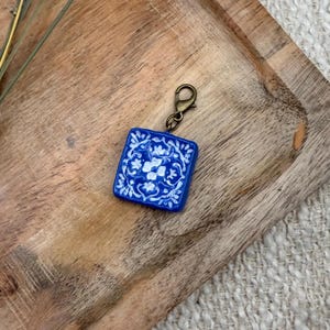 May include: A square charm featuring a deep blue background with a white floral and scrollwork pattern. The charm is attached to a bronze-toned metal lobster clasp, providing a secure attachment point for various accessories or jewelry items.