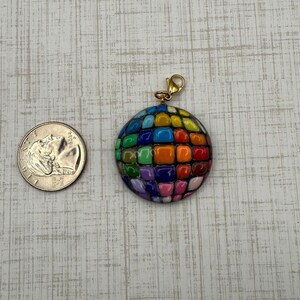 Puede incluir: Un colgante redondo y colorido con un cierre dorado. El colgante est&aacute; hecho de peque&ntilde;as baldosas cuadradas en varios colores, incluyendo rojo, naranja, amarillo, verde, azul y morado. Una moneda est&aacute; al lado del colgante para la comparaci&oacute;n de tama&ntilde;os.