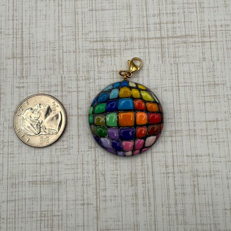 Puede incluir: Un colgante redondo y colorido con un cierre dorado. El colgante est&aacute; hecho de peque&ntilde;as baldosas cuadradas en varios colores, incluyendo rojo, naranja, amarillo, verde, azul y morado. Una moneda est&aacute; al lado del colgante para la comparaci&oacute;n de tama&ntilde;os.