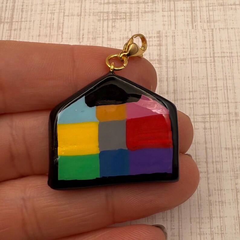 May include: A colorful house charm with a gold-colored clasp. The house is divided into nine squares, each painted a different color, including blue, yellow, green, red, and purple. The charm has a black outline.