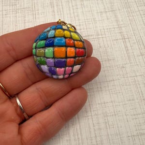 Puede incluir: Un colgante esf&eacute;rico de estilo mosaico, con una anilla dorada. La superficie del colgante est&aacute; compuesta por peque&ntilde;as baldosas cuadradas de varios colores, como azul, verde, naranja y rosa. El colgante est&aacute; sujeto en una mano.
