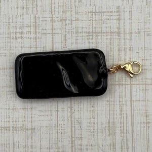 May include: A rectangular, glossy black charm with rounded corners. A small gold-colored lobster clasp is attached to the charm, ready to be added to a necklace or bracelet. The charm is a solid, opaque color.