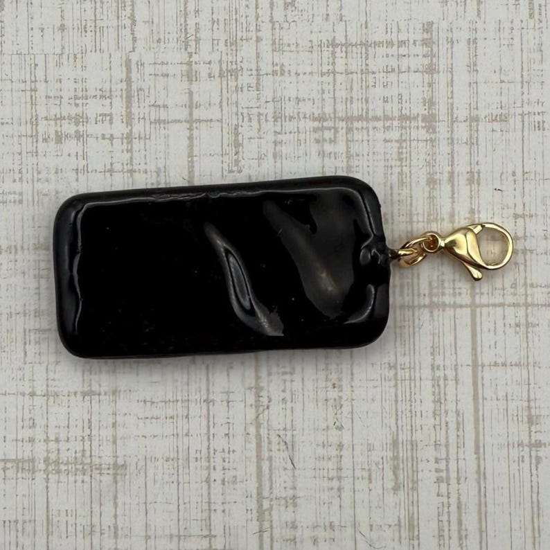 May include: A rectangular, glossy black charm with rounded corners. A small gold-colored lobster clasp is attached to the charm, ready to be added to a necklace or bracelet. The charm is a solid, opaque color.