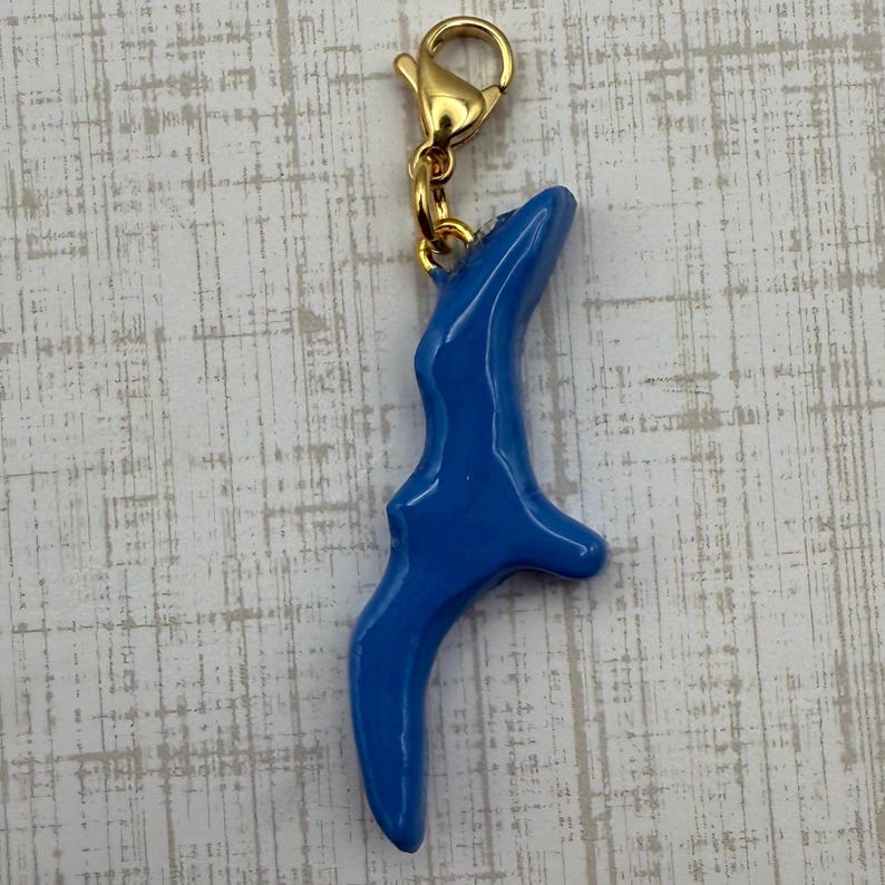 May include: A blue charm with a gold-colored clasp. The charm has a curved, abstract shape. The charm is a bright, solid blue color and appears to be made of a smooth material.