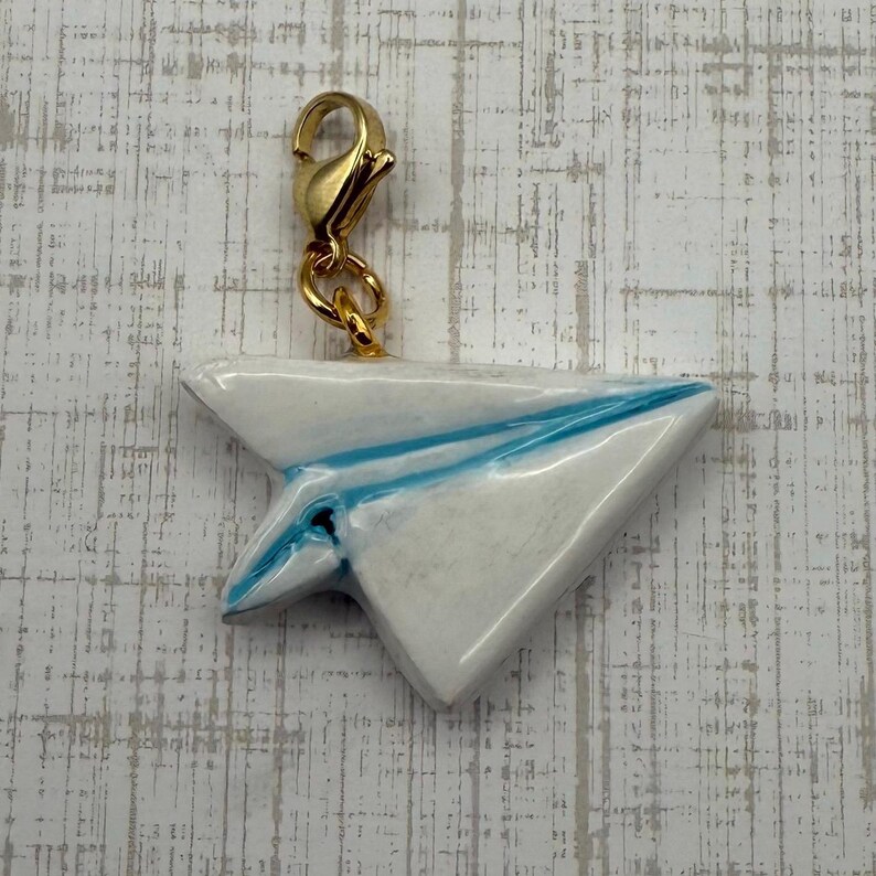 May include: A white paper airplane charm with blue accents and a gold-colored clasp. The charm is a miniature replica of a paper airplane, with blue lines mimicking the folds. The clasp is attached to the top of the airplane.