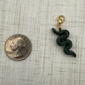 May include: A dark green snake charm with a gold-colored clasp. The charm is next to a U.S. quarter coin for size comparison. The snake charm is approximately 1 inch in length.