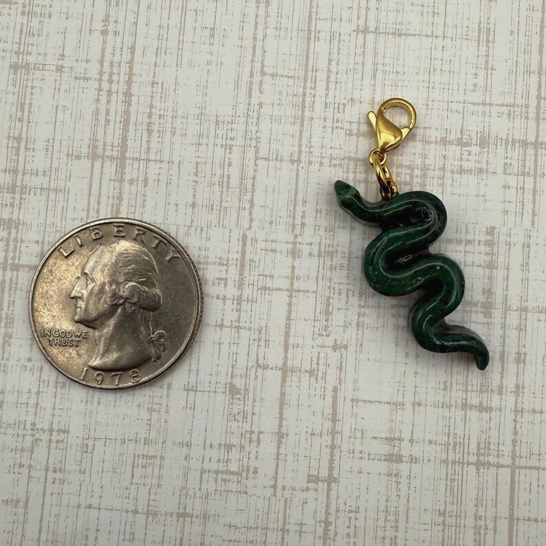 May include: A dark green snake charm with a gold-colored clasp. The charm is next to a U.S. quarter coin for size comparison. The snake charm is approximately 1 inch in length.