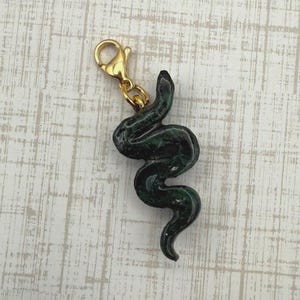 May include: A dark green snake charm with a gold-colored clasp. The snake is sculpted with a textured surface and a curved, serpentine design. The charm is approximately 2 inches long.
