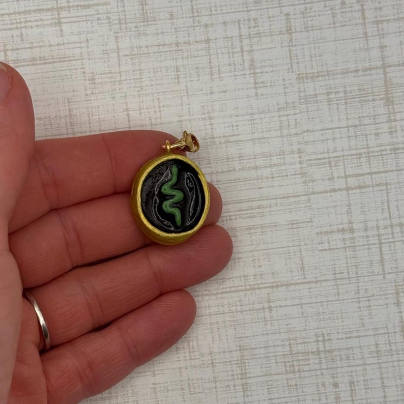 May include: Gold-colored pendant with a black background and a green snake design. The pendant is round with a gold-colored border and a small loop for a chain. The snake is a stylized, wavy design.
