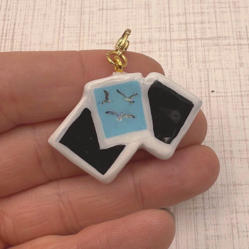 May include: A gold-colored charm featuring three miniature polaroid-style images. One image has a light blue background with three seagulls. The other two images are black. The charm is attached to a gold-colored clasp.