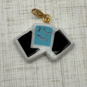 May include: A charm with a gold-colored clasp featuring a design of three seagulls flying over a light blue background. The charm has a white border with black accents, resembling a photograph.