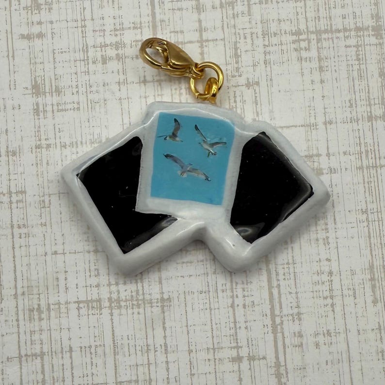 May include: A charm with a gold-colored clasp featuring a design of three seagulls flying over a light blue background. The charm has a white border with black accents, resembling a photograph.