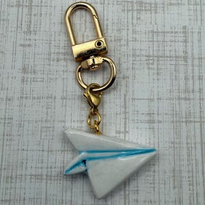 May include: A gold-colored keychain with a white paper airplane charm. The paper airplane has blue accents and is attached to the keychain with a gold-colored clasp and ring. The background is a light gray and white wood grain pattern.
