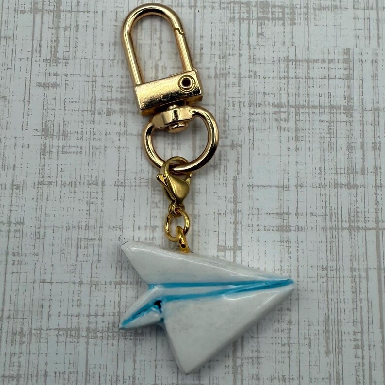 May include: A gold-colored keychain with a white paper airplane charm. The paper airplane has blue accents and is attached to the keychain with a gold-colored clasp and ring. The background is a light gray and white wood grain pattern.
