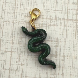 May include: A dark green snake charm with a gold-colored clasp. The snake is detailed with a smooth, glossy finish. The charm is designed to be attached to a bracelet or necklace. The background is a light, textured surface.