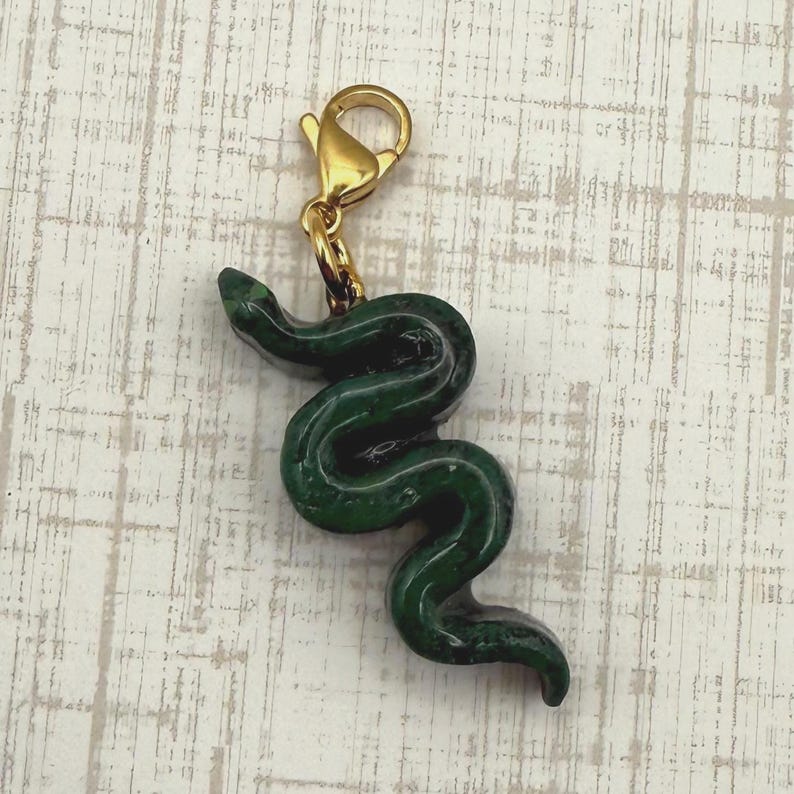 May include: A dark green snake charm with a gold-colored clasp. The snake is detailed with a smooth, glossy finish. The charm is designed to be attached to a bracelet or necklace. The background is a light, textured surface.